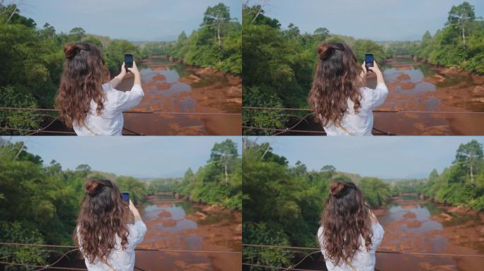 女子站在桥上，用智能手机拍摄热带河流