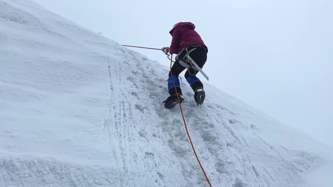 一名女登山者在垂直栏杆上的竹马的帮助下爬上陡峭的冰雪斜坡。