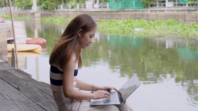 亚洲女孩坐在运河悬挂腿与自然的气氛，微风，坐着工作与膝上电脑在运河悬挂的腿，在笔记本电脑上打字找地方
