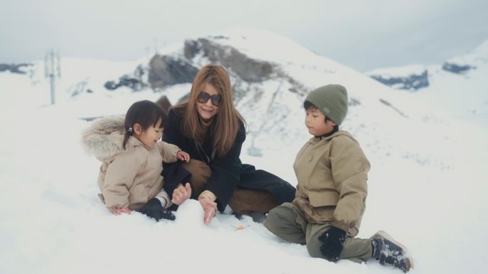 快乐的妈妈和她的两个孩子，坐在雪地上，一起玩堆雪人作为团队在冬天。