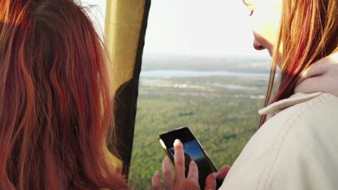 两个女孩乘坐热气球飞行，其中一个给另一个看她用智能手机拍的人像照片