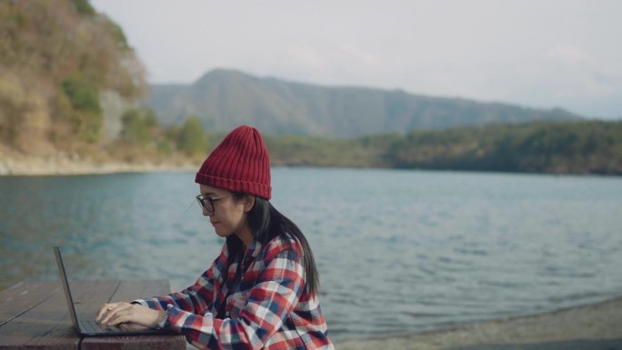 一个女人穿着红黑相间的格子衬衫，戴着红色的针织帽坐在湖边，她在用笔记本电脑给她的客户发电子邮件