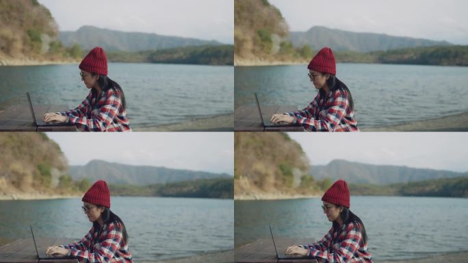 一个女人穿着红黑相间的格子衬衫，戴着红色的针织帽坐在湖边，她在用笔记本电脑给她的客户发电子邮件