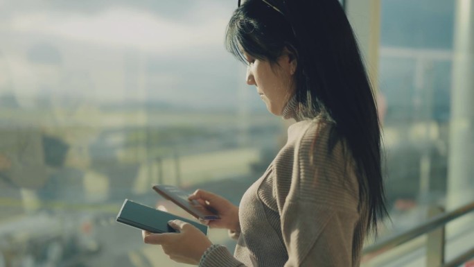 一名亚洲女子站在机场窗口观察天空，在智能手机上查看护照和时间