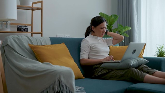 一位亚洲女性坐在家里客厅的沙发上，用笔记本电脑忍受着疼痛，伸展和按摩自己的身体。