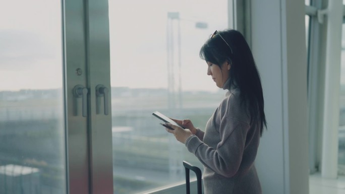 一名亚洲女子站在机场透过窗户观察天空，看护照，在智能手机上查看时间