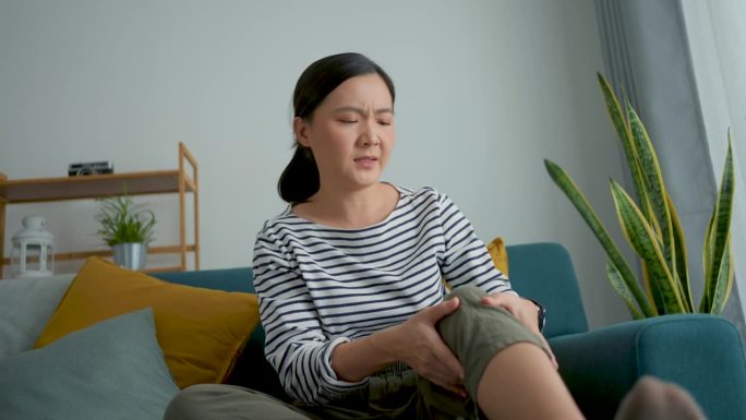 亚洲妇女坐在家里客厅的沙发上感到膝盖受伤疼痛。