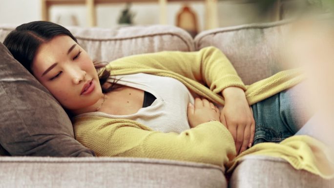 疼痛，经期和亚洲妇女用手放在肚子上怀孕，月经或腹部痉挛。健康，消化问题和女孩在沙发上抱着腹部子宫内膜