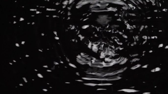 俯视图:黑色背景上孤立的水滴落在水面上，夜晚雨滴落在地面上时的混沌波型慢镜头