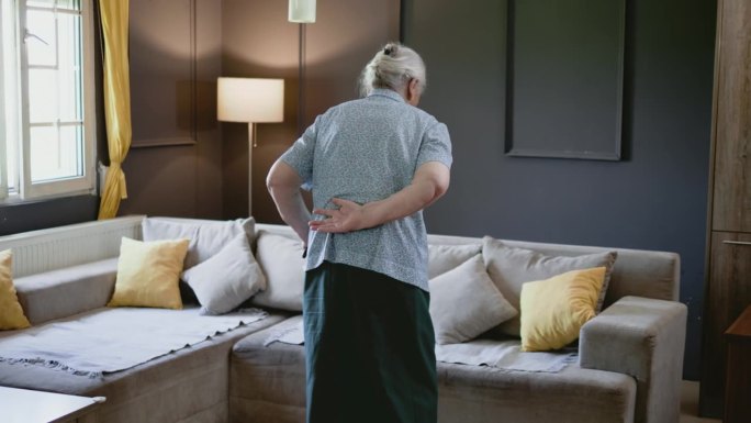 80年代退休的老妇人拄着拐杖试图坐在客厅的沙发上。这位老人走路很吃力。膝关节和关节疼痛