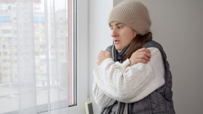 戴着帽子，穿着毛衣的年轻女子在家里看着窗外，冻得要命。能源危机的概念，高账单，坏了的供暖系统，节约和