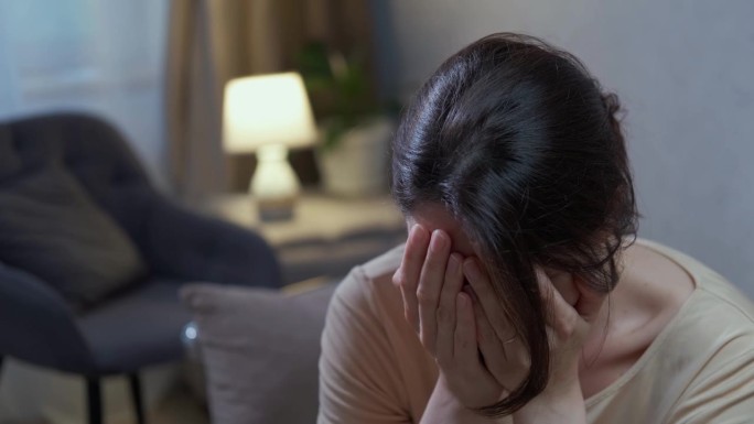 一个白人女人坐在沙发上哭，用手捂着脸，沮丧地坐在房间的沙发上。精神健康问题，虐待