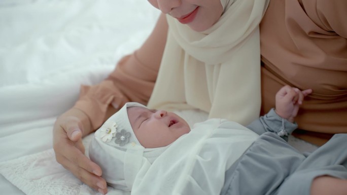 让她的女儿不哭，快乐的穆斯林母亲戴着淡黄色的头巾，躺在卧室的白色床上照顾她，让她快乐