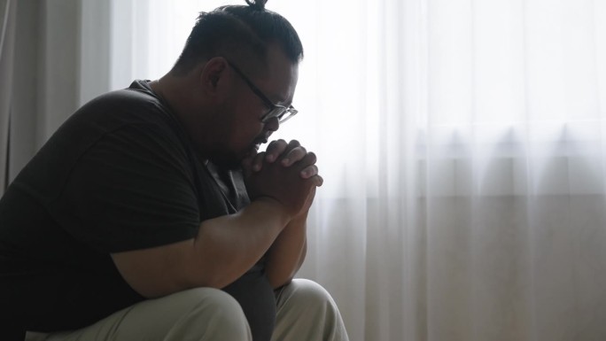 侧视图:一个胖乎乎的亚洲人不安地坐在客厅的沙发上，低着头，双手交叉，祈祷奇迹发生在他身上。
