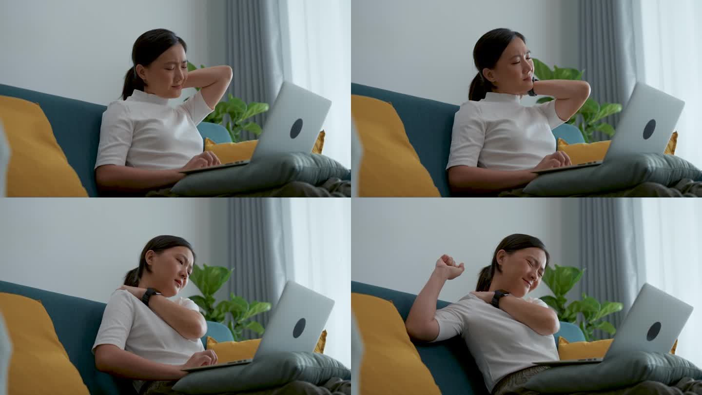 一位亚洲女性坐在家里客厅的沙发上，用笔记本电脑忍受着疼痛，伸展和按摩自己的身体。