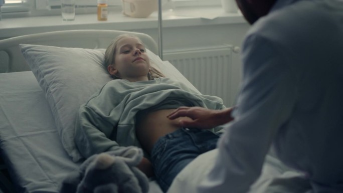 医生在病房里触诊女孩的胃痛。生病的孩子躺在医院的病床上。