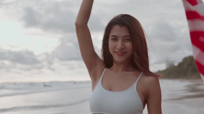 7月4日独立纪念日，年轻的亚洲女子在海滩上享受时光，在日落的天空下，在夏天的海滩上挥舞着美国国旗。