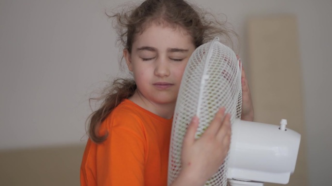 孩子在家里享受电风扇吹来的凉风。女孩在通风机前遭受高温折磨，用电风扇冷却器给自己降温。炎热气候变化。