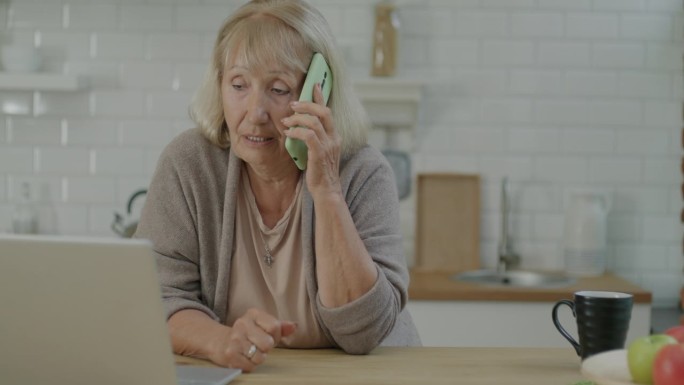 老年妇女在家里的厨房里打电话和使用笔记本电脑