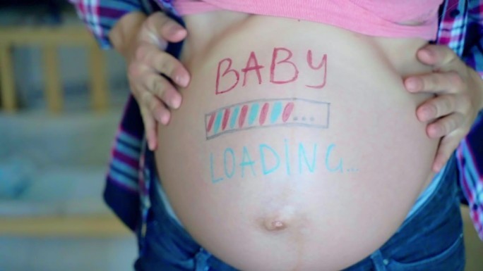 蓝粉相间的婴儿在孕妇的肚子上写着铭文，期待着妈妈等待孩子的到来，快乐的女性慢慢地在镜头前摆姿势，竖起
