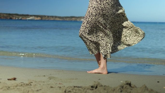 不认识的女人光着脚，穿着雪纺裙在海风中飘动。女腿赤着脚站在沙滩海岸线上，轻风摇曳长裙。