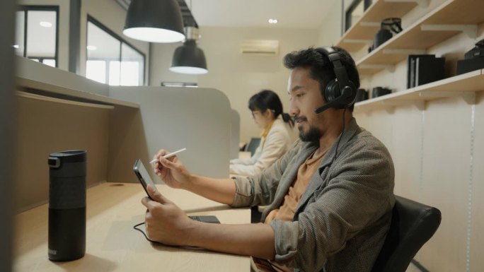 亚洲男人看起来很专注。他使用平板电脑，在工作空间做一些笔记