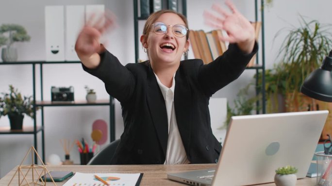 在笔记本电脑上工作的女商人对着镜头友好地微笑，在办公室里挥手打招呼