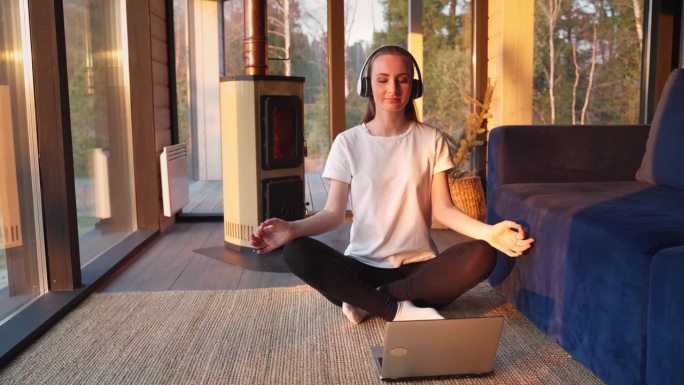 年轻女子戴着耳机进行在线瑜伽冥想练习。一名女子在客厅里以壁炉为背景，在笔记本电脑显示器前坐成莲花状，