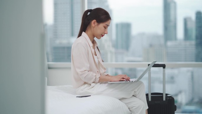 亚洲女商人用科技笔记本电脑在床上工作，而在市中心带着行李箱进入房间或办公室