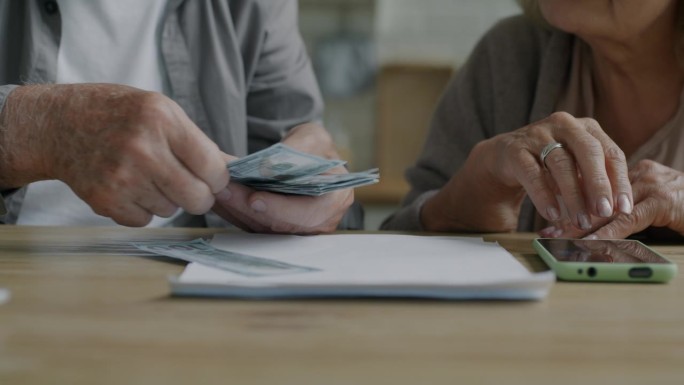 老年夫妇的手特写，在家里的桌子上用智能手机计算付款，拿着钱