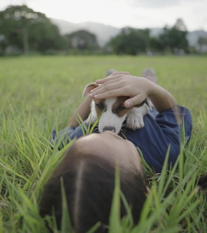一个亚洲女孩躺在公共自然公园里，她的小狗趴在她的身上，抚摸着她的狗