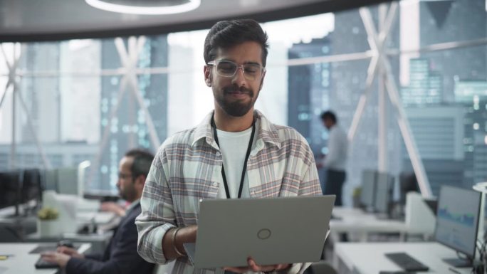 英俊的印度男子戴着眼镜，迷人地微笑着看着镜头。年轻聪明的男工程师拿着笔记本电脑。现代企业办公室的财务