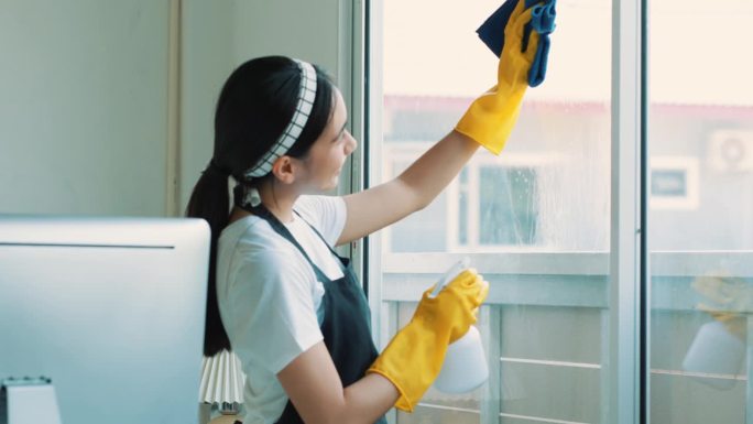一个打扫房子的女人的特写镜头。她一边打扫桌子，一边用喷雾器和蓝色布擦灰尘。房屋清洁概念。