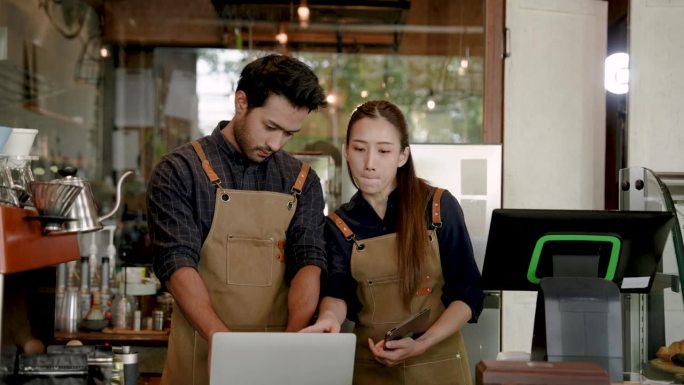 漂亮的亚洲女人和印度男人小企业主夫妇看着从电脑屏幕进入咖啡馆的订单。以及通过在线应用程序销售的平板电