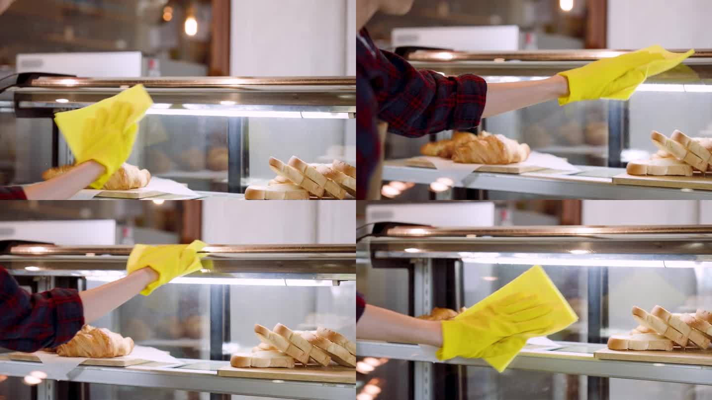 近距离的玻璃柜中拿着咖啡面包店工人的手，清洁玻璃柜中使用的面包店物品，有玻璃清洁剂在手喷在玻璃上柔软