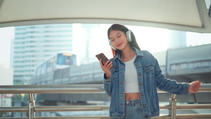 快乐的年轻亚洲女子戴着耳机在城市的街道上欣赏音乐，微笑的女性独自听音乐跳舞。