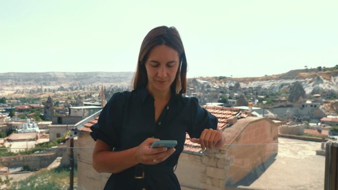 年轻的女游客在度假时用智能手机远程工作，看着身后美丽的景色