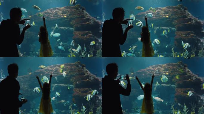爸爸用智能手机给女儿拍照，小女孩看着鱼儿在珊瑚礁里游泳，爸爸在社交媒体上分享海洋馆的乐趣