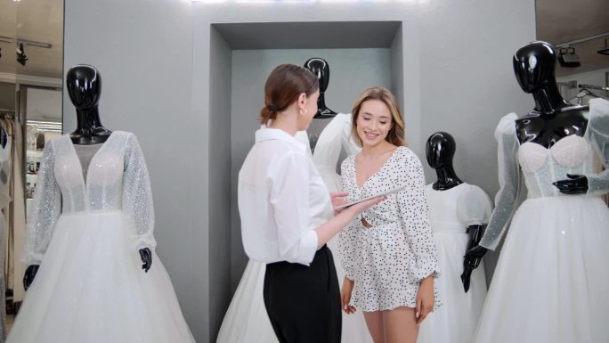 有吸引力的新娘店老板设计师女士用平板电脑帮助和支持顾客在商店选择婚纱