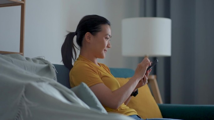亚洲女人坐在客厅的沙发上用智能手机开灯。