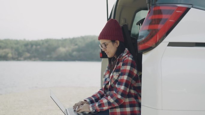 一名亚洲年轻女子在一辆停在乡下湖边的汽车后座上使用她的笔记本电脑。