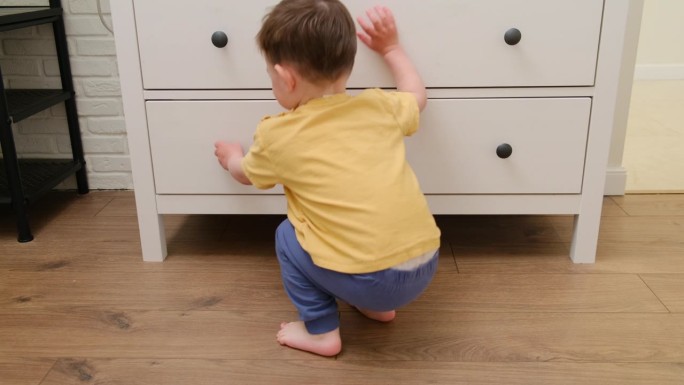 一个小男孩正在玩一个打开的梳妆台抽屉，没有意识到潜在的危险。室内游乐区的柜子对婴儿安全构成威胁。大约