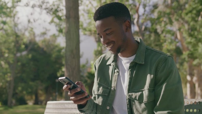 快乐的年轻黑人穿着衬衫在公园树下的长椅上玩手机