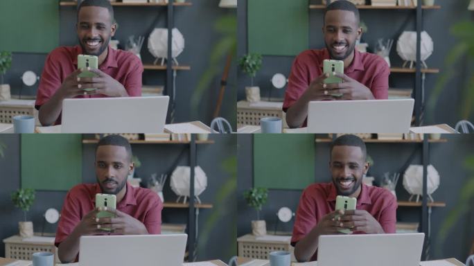 开朗的非洲裔美国商人看着智能手机屏幕，微笑着坐在办公室的办公桌前
