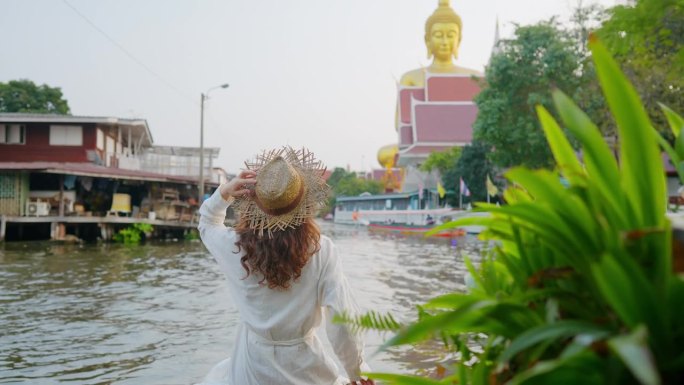 一名女子坐在曼谷的河岸上，看着巨大的金佛雕像