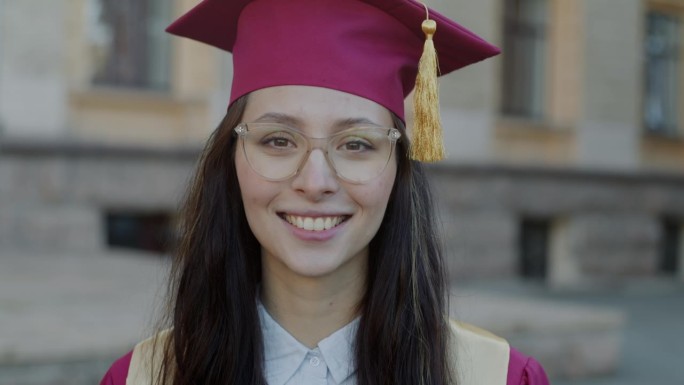 快乐的年轻女大学毕业生的肖像，戴着帽子，穿着长袍，在校园里微笑