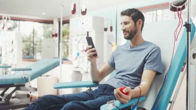 白人男子在医院为有需要的人献血。男性献血者挤压心形红球泵血，用智能手机在线聊天。为意外事故受害者捐款
