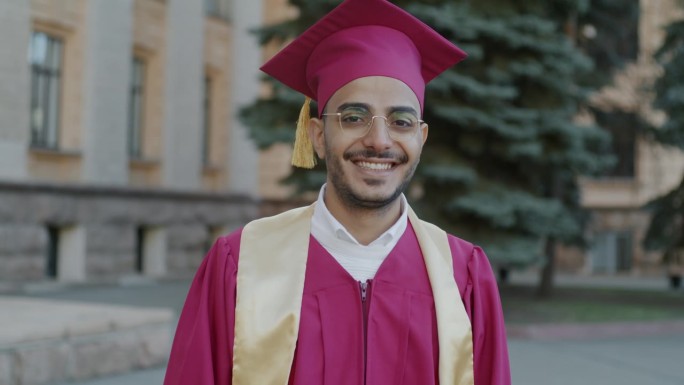 慢动作肖像的阿拉伯男子毕业生站在校园里穿着长袍和砂浆板微笑
