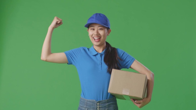 亚洲女快递员穿着蓝色制服，微笑着，肌肉发达，在工作室绿屏背景下送纸箱