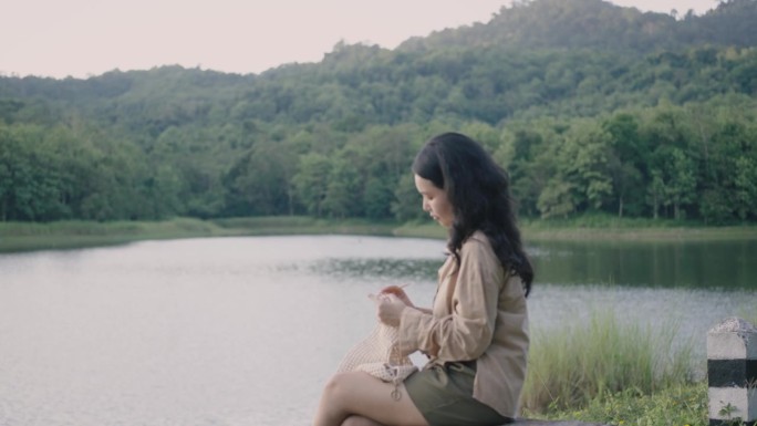 一个阳光明媚的夏日，一位亚洲年轻女子正在河边用奶油色的纱线编织。